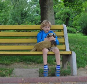 little boy with blond hair in shorts and blue jacket sitting alone on bench holding stuffed animal